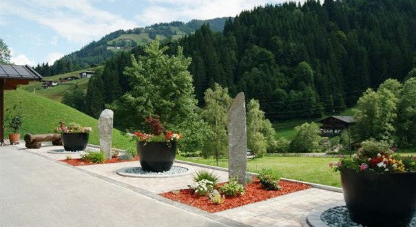 Pflanzkübel Fiberglas rund konisch D110xH95cm elegant schwarz-matt als Objektbepflanzung mit Blumen an einer Einfahrt vor bewaldeter Berglandschaft