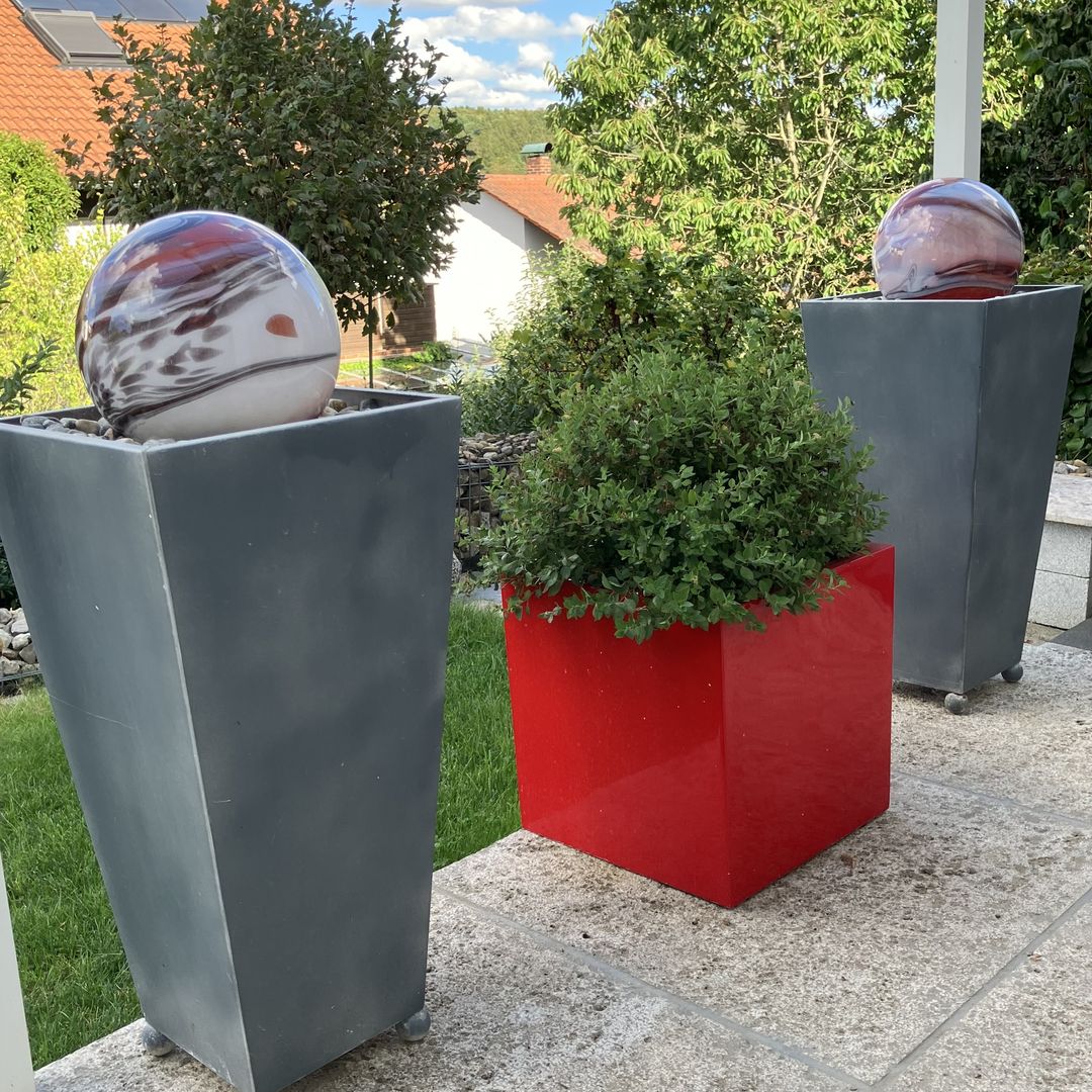 Pflanzkübel Fiberglas quadratisch 38x38x38cm hochglanz rot mit Spierstrauch auf einer Terrasse, daneben zwei hohe graue Pflanzsäulen