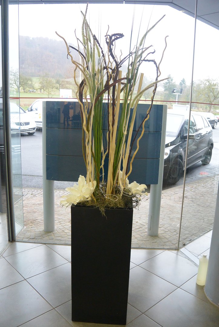Pflanzkübel Fiberglas Säule 38x38x100cm elegant schwarz matt mit einem Gesteck aus Ziergräsern und weißen Orchideenblüten in einem verglasten Eingangsbereich