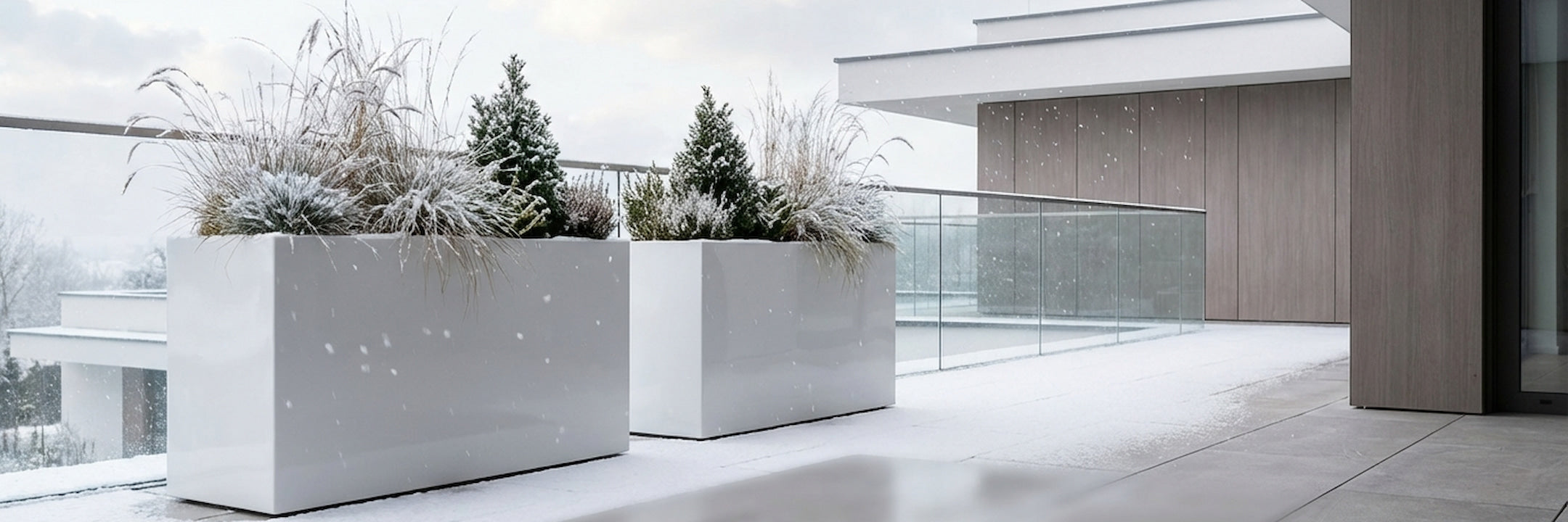 Hochglanz weiße Fiberglas-Pflanzkübel mit Ziergräsern und kleinen Koniferen auf verschneiter Terrasse – moderne Pflanzgefäße